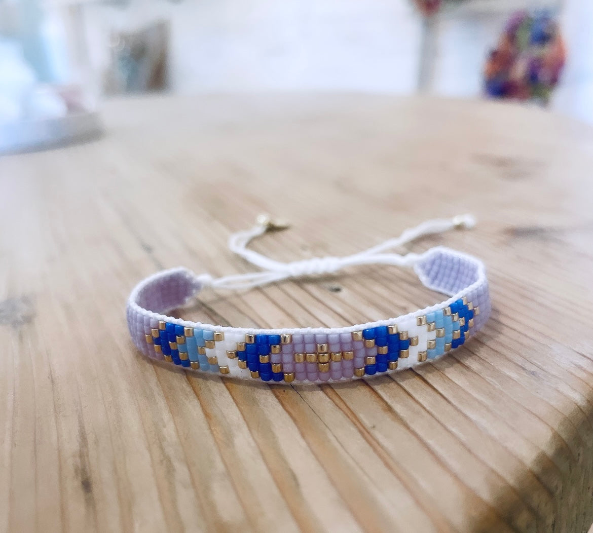 Gewebtes Armband in der Farbe weiß/lila/flieder/hell blau & gold. Durch den Schiebeknoten ist es variabel verstellbar. Mit Miyuki Glasperlen