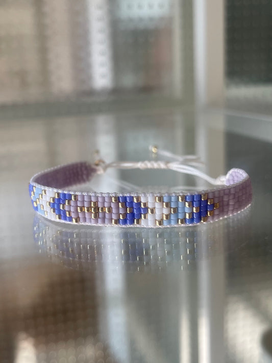 Gewebtes Armband in der Farbe weiß/lila/flieder/hell blau & gold. Durch den Schiebeknoten ist es variabel verstellbar. Mit Miyuki Glasperlen