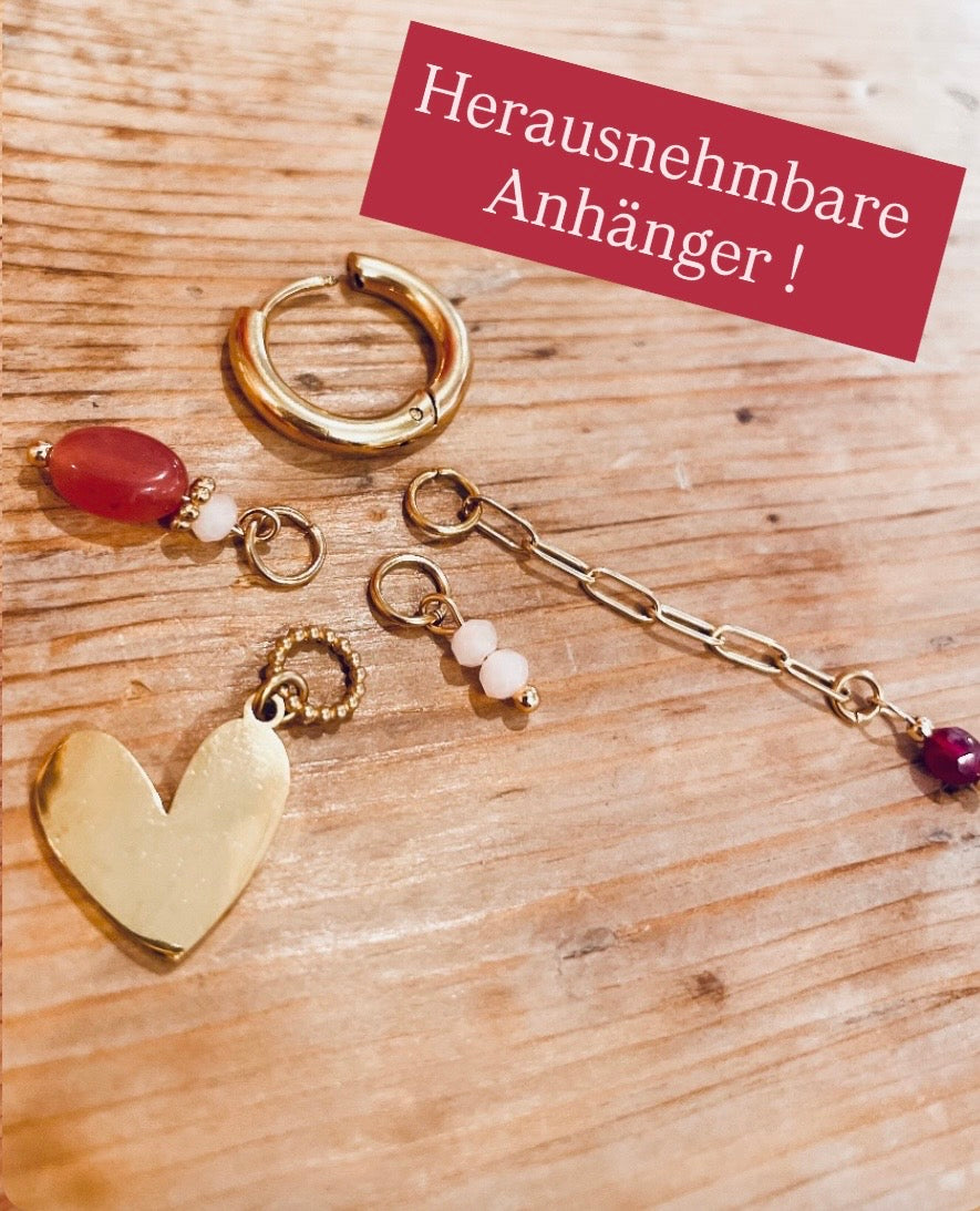 In goldene Click Creole ist ein gedrehter Ring eingehangen mit poliertem Herzchen. Zudem sind weitere kleine rosa Steinchen eingehangen und ein kleines Edelstahlkettchen