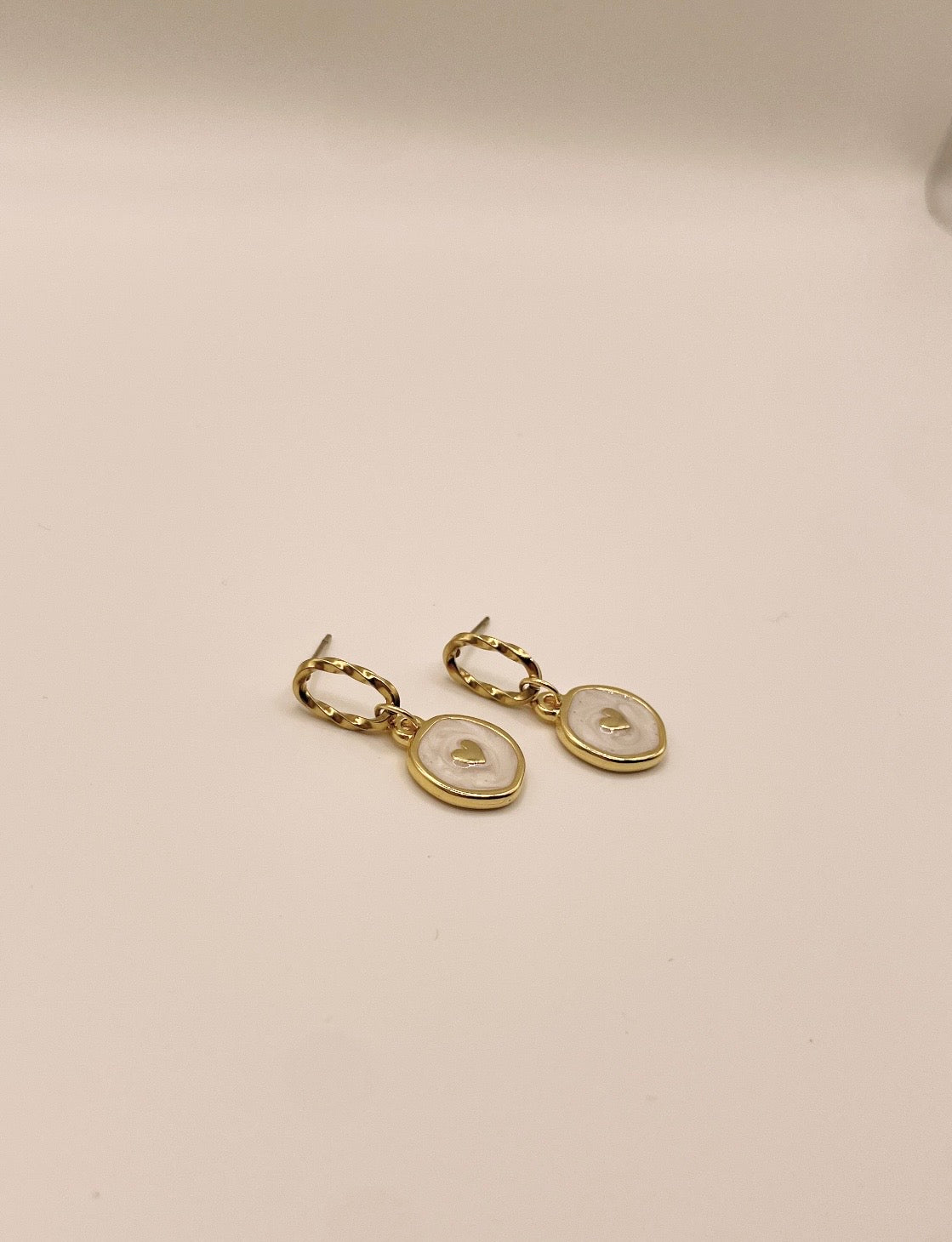 Ovaler gedrehter Edelstahlstecker mit eingehängtem runden Anhänger. Der Anhänger ist mit weißer Perlmutt Emaille bemalt und einem goldenen Edelstahlherz.