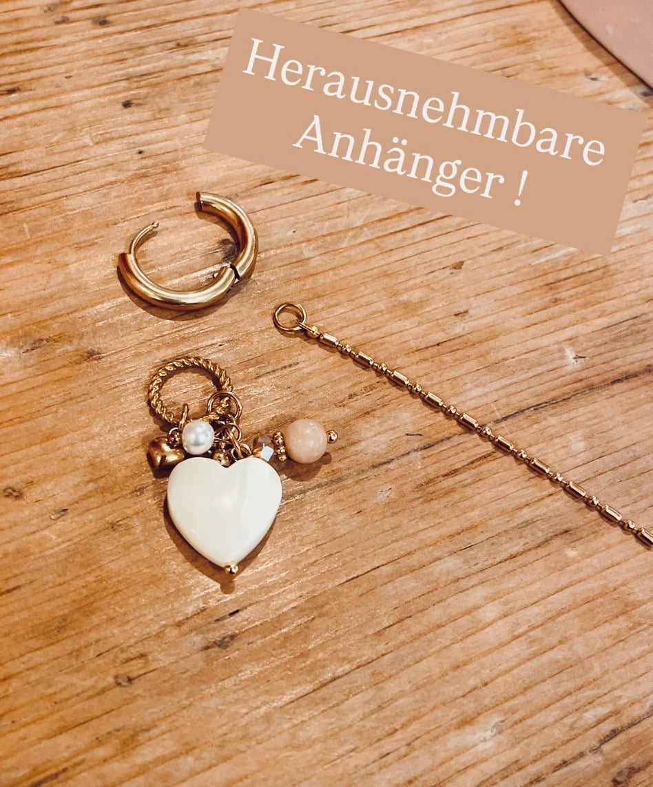 In die Goldene Click Creole ist ein gedrehter Ring eingehangen. An dem Ring hängt ein weißes Perlmutt Herz. Zudem sind weitere kleine Steinchen in nude und weiß